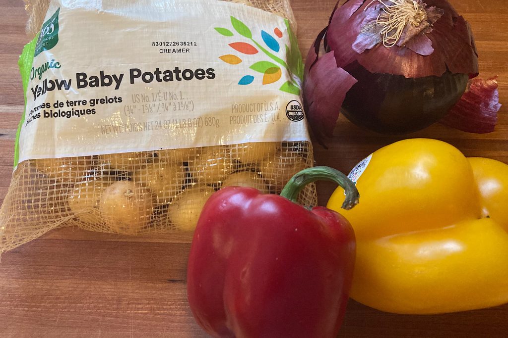 Bag of baby yellow potatoes, a red and yellow bell pepper and a red onion on a wooden cutting board