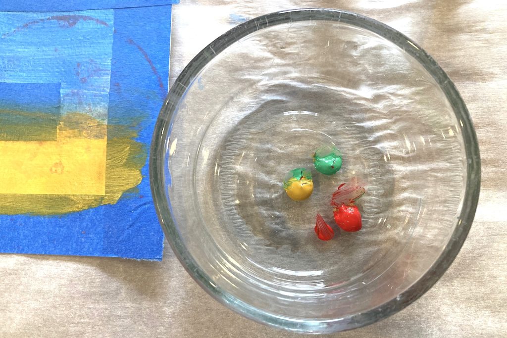 green, yellow and red acrylic paint drops in glass bowl next to taped off artwork